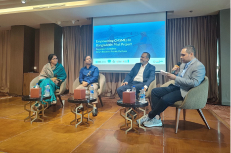 Four individuals engage in a panel discussion on a well-lit stage. A projector screen displays information about the ‘Empowering MSMEs in Bangladesh Pilot Project.’